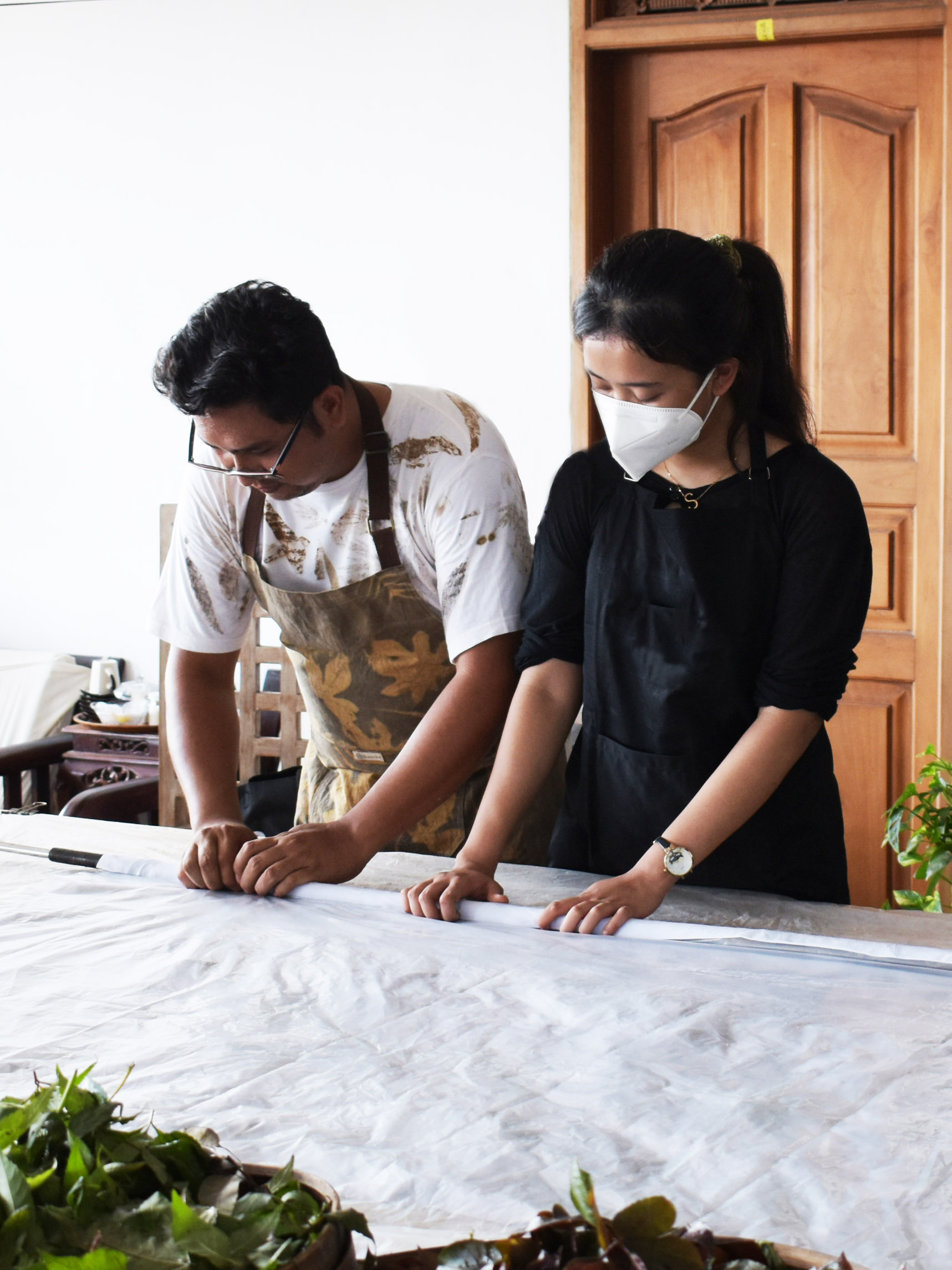 Two people working together on a project, one wearing a mask and apron, the other wearing a mask and black clothing.